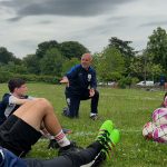 A coach kneels on grass to talk to teenage footballers