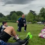 A coach kneels on grass to talk to teenage footballers