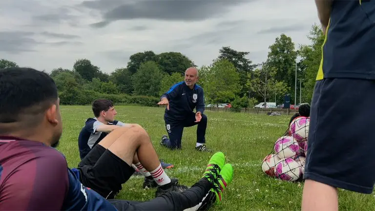 A coach kneels on grass to talk to teenage footballers