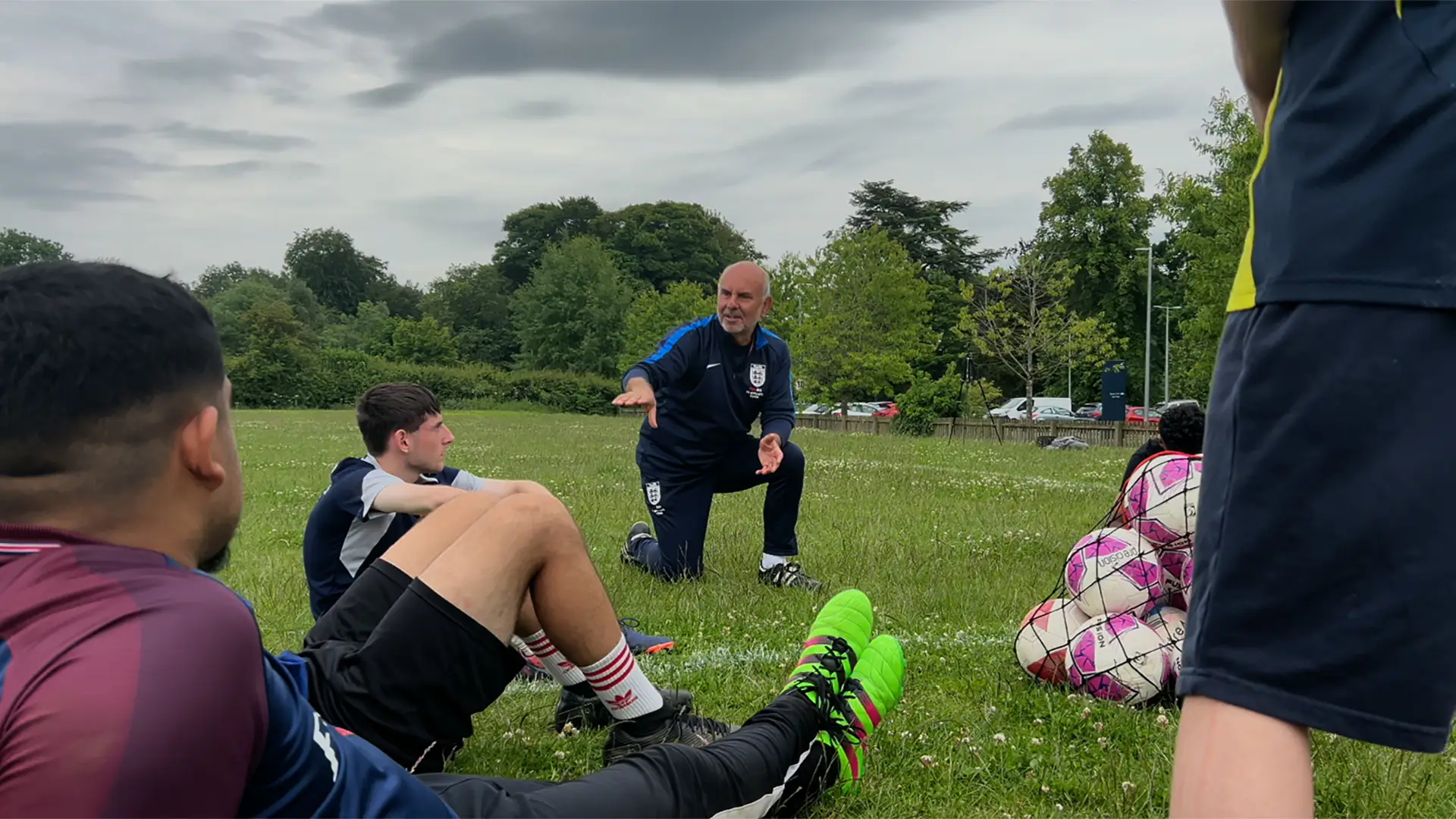 A coach kneels on grass to talk to teenage footballers