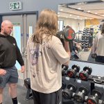 A young man faces a mirror and lifts a weight. He is watched by a coach.