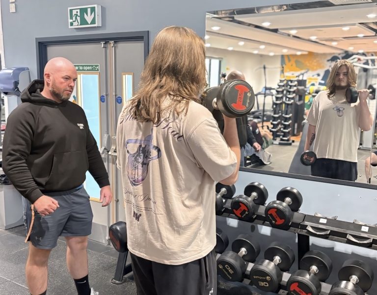 A young man faces a mirror and lifts a weight. He is watched by a coach.