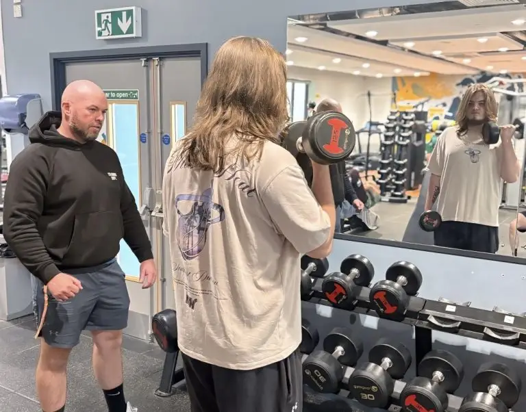 A young man faces a mirror and lifts a weight. He is watched by a coach.