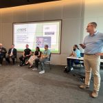 Spencer Moore stands beside a seated group of experts, leading a presentation