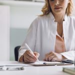 A doctor sits at a desk, writing on a clipboard