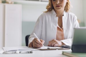 A doctor sits at a desk, writing on a clipboard