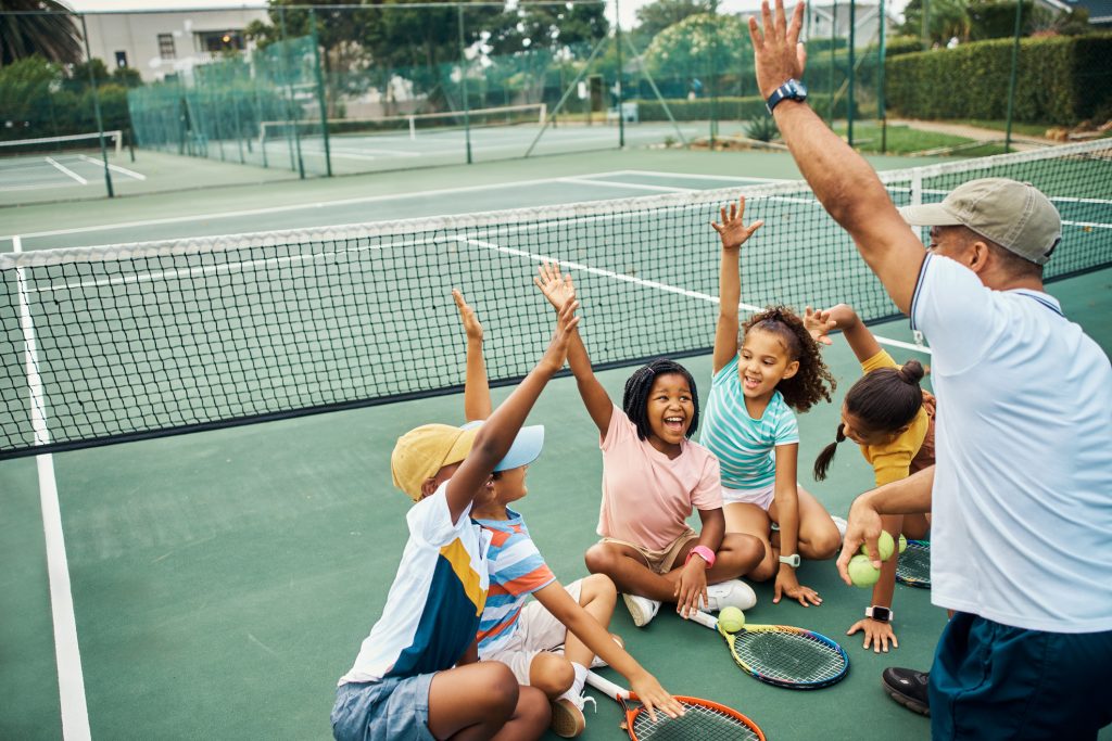 Children sit on the floor around a coach. They all have one hand in the air and are smiling