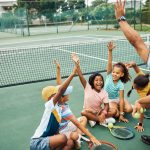 Children sit on the floor around a coach. They all have one hand in the air and are smiling
