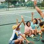 Children sit on the floor around a coach. They all have one hand in the air and are smiling