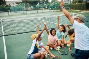 Children sit on the floor around a coach. They all have one hand in the air and are smiling