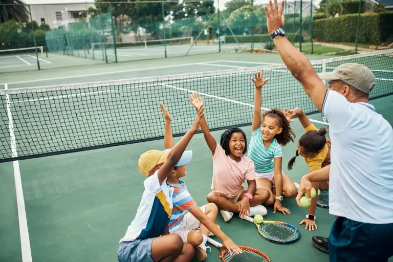 Children sit on the floor around a coach. They all have one hand in the air and are smiling