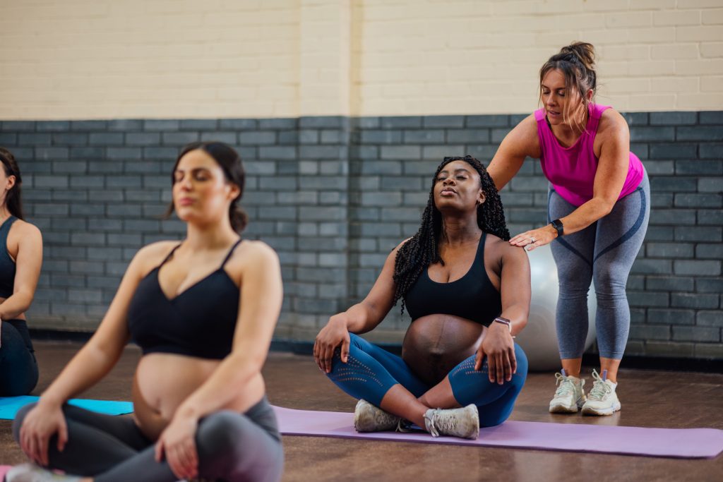 A yoga teacher touches the shoulders of a pregnant student sitting cross-legged on a mat.