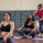 A yoga teacher touches the shoulders of a pregnant student sitting cross-legged on a mat.