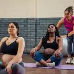 A yoga teacher touches the shoulders of a pregnant student sitting cross-legged on a mat.