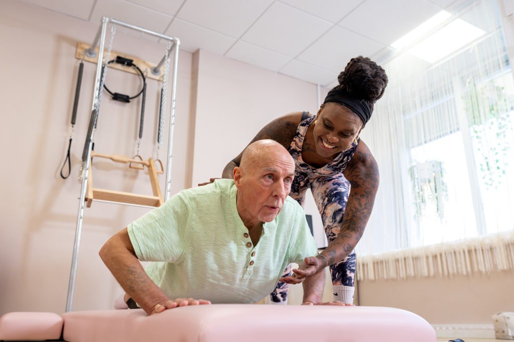 A female Pilates instructor supports an older male client in a plank position.