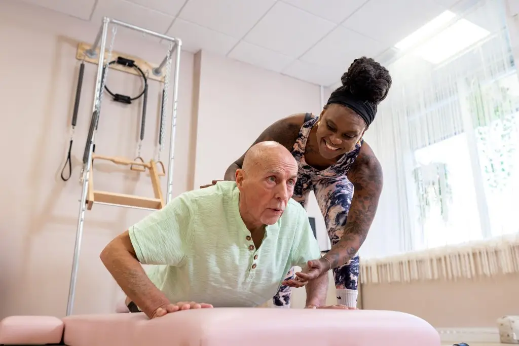 A female Pilates instructor supports an older male client in a plank position.