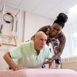 A female Pilates instructor supports an older male client in a plank position.