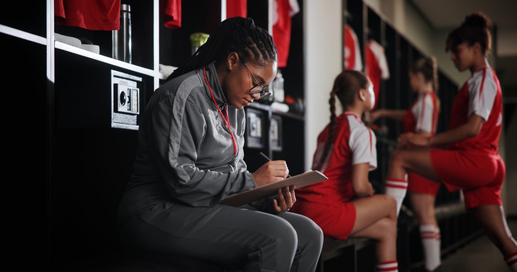 A female coach sits in a locker room focusing on a clipboard