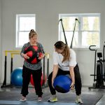 Two women stand in a gym picking up medicine balls