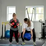 Two women stand in a gym picking up medicine balls