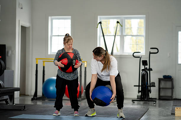 Two women stand in a gym picking up medicine balls