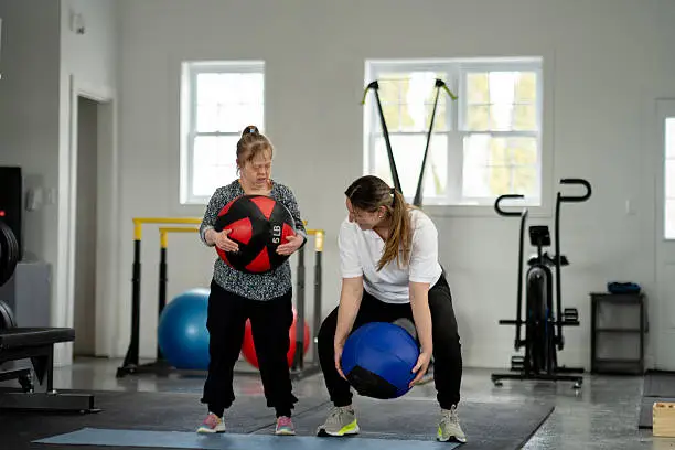 Two women stand in a gym picking up medicine balls