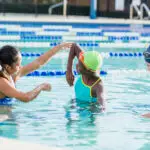 A young woman and three children are standing in a pool. The woman is instructing the children on swimming technique.