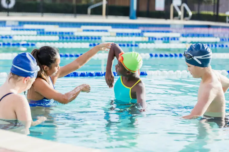 A young woman and three children are standing in a pool. The woman is instructing the children on swimming technique.