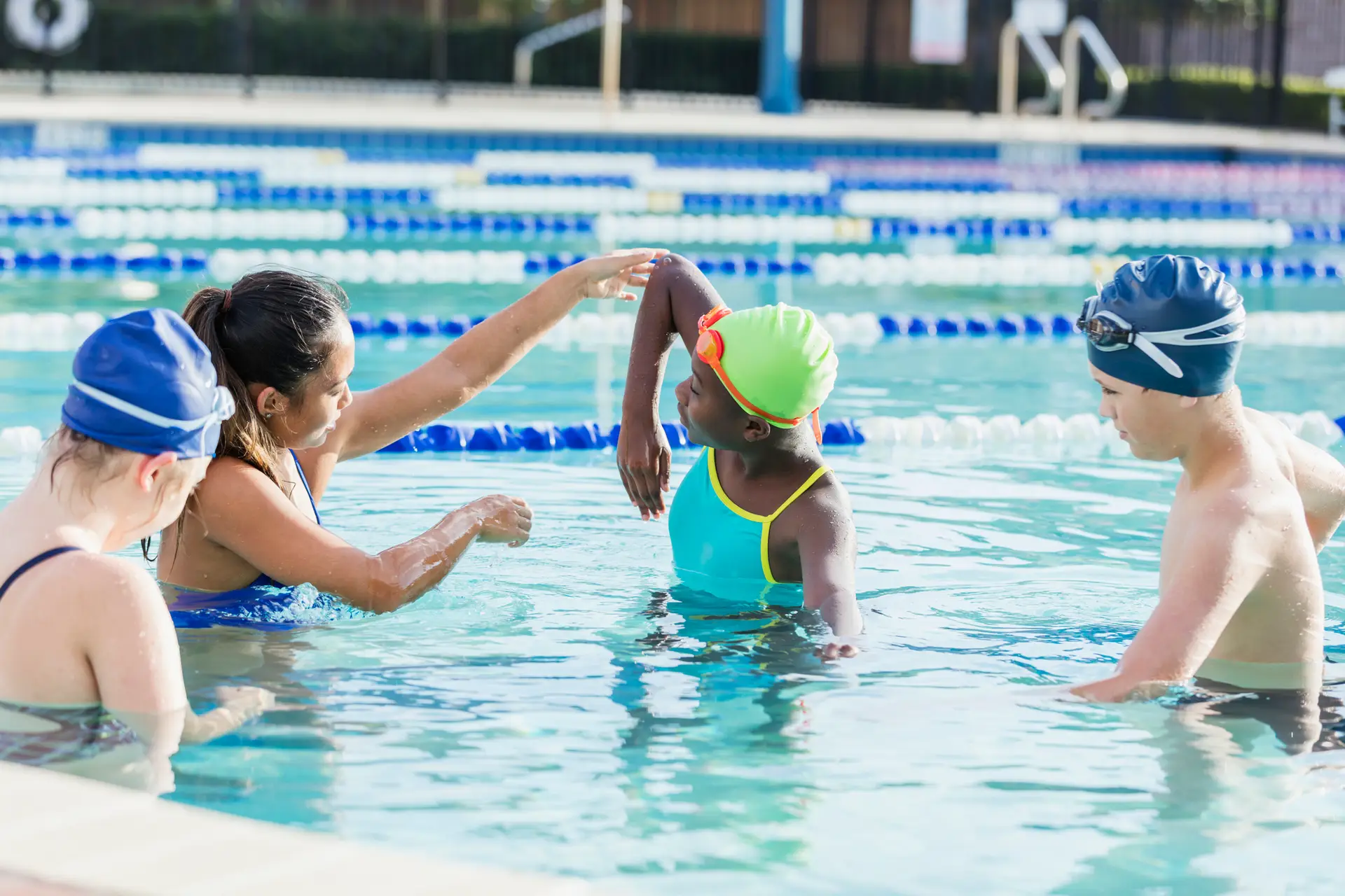 A young woman and three children are standing in a pool. The woman is instructing the children on swimming technique.