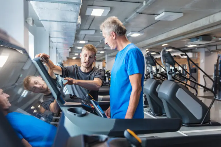 A personal trainer guides a client using a treadmill, pointing at the screen
