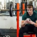 A young man sits on a gym bench with headphones on looking at his phone.