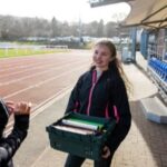 Two female teenagers standing and talking at an outdoor running track. One of the girls is holding a crate of relay batons.