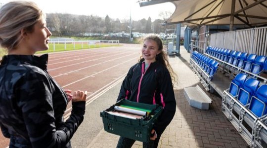 Two female teenagers standing and talking at an outdoor running track. One of the girls is holding a crate of relay batons.