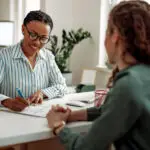 Two women sit either side of a desk in an office. One makes notes on paper.
