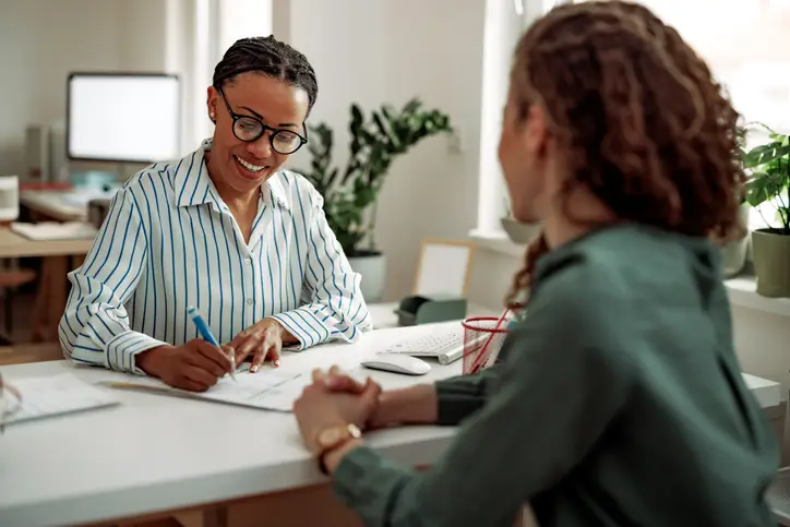 Two women sit either side of a desk in an office. One makes notes on paper.