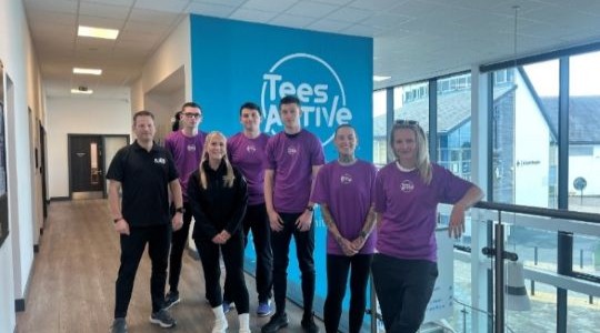 A group of young adults stand in front of a Tees Active logo in a leisure centre