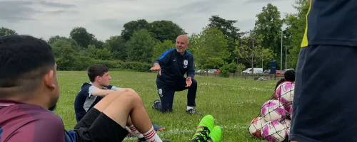 A coach kneels on grass to talk to teenage footballers
