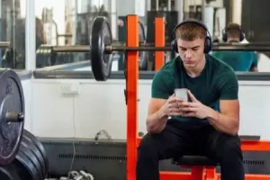 A young man sits on a gym bench with headphones on looking at his phone.