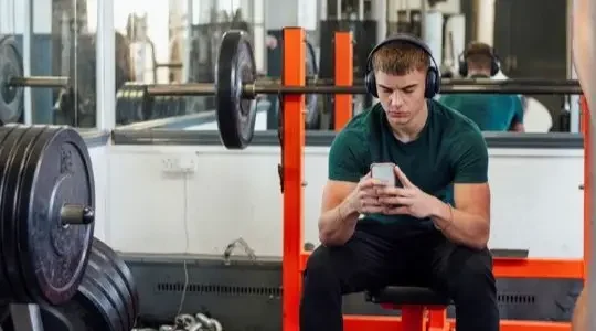 A young man sits on a gym bench with headphones on looking at his phone.