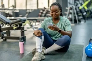 A woman sits on the floor of a gym looking at her phone