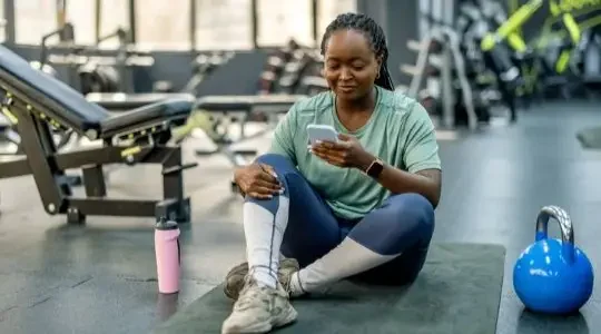 A woman sits on the floor of a gym looking at her phone